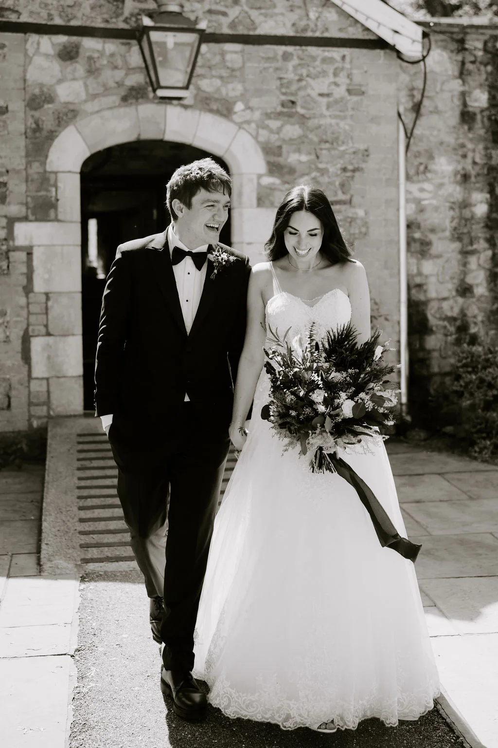 Pencoed House Estate Wedding Photography | Cardiff Amy and Dan walking hand-in-hand beneath the historic stone archway at Pencoed House Estate in Cardiff