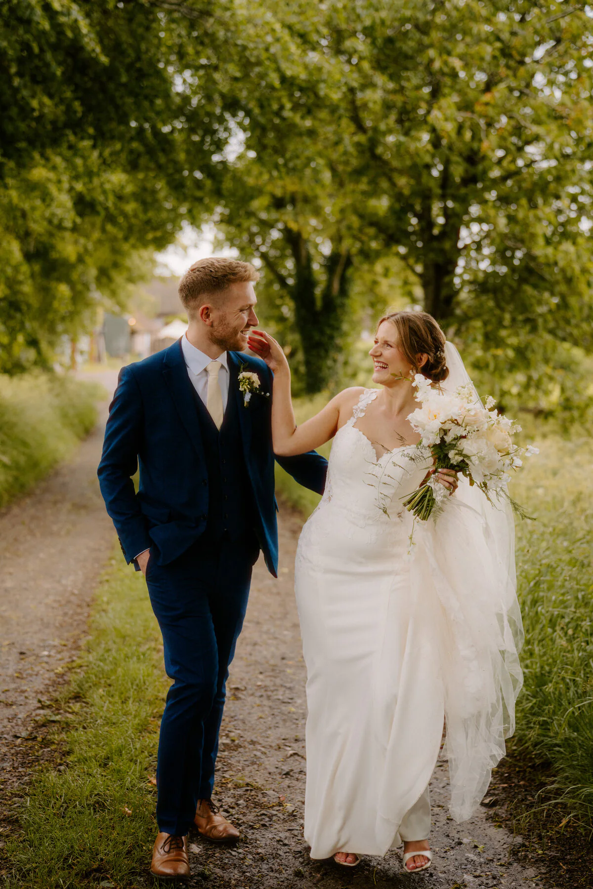 Ca102481 Edit Bride and groom laughing as they walk hand in hand down a tree-lined avenue.