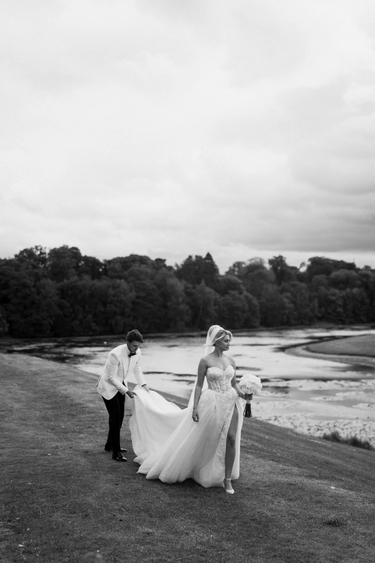 Ca100310 Black and white photograph of a bride walking outdoors while her dress is carried.