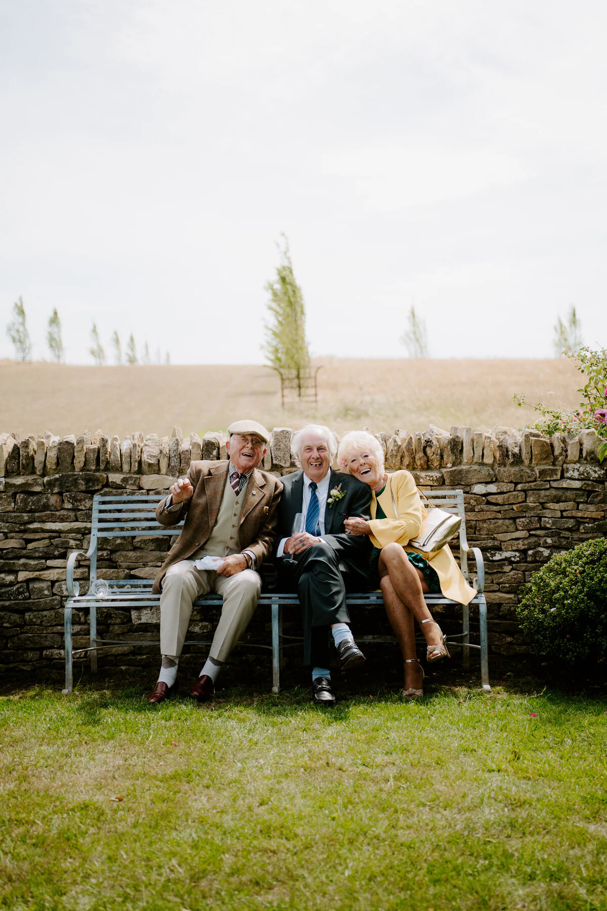 Dsc07033 Three wedding guests laughing together on a bench beside a stone wall.