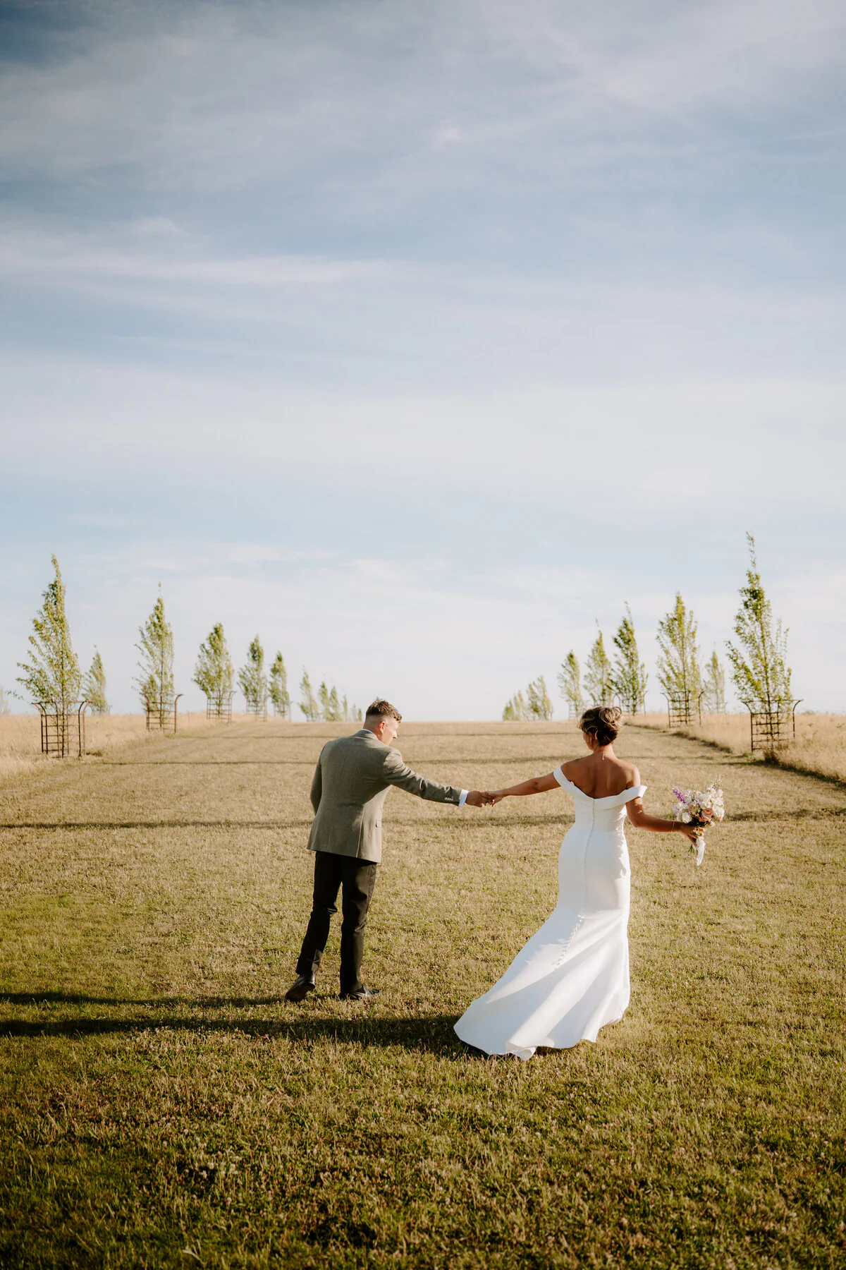 Dsc09196 Bride in an off-shoulder gown and groom in grey tweed walking up a grassy hillside path.
