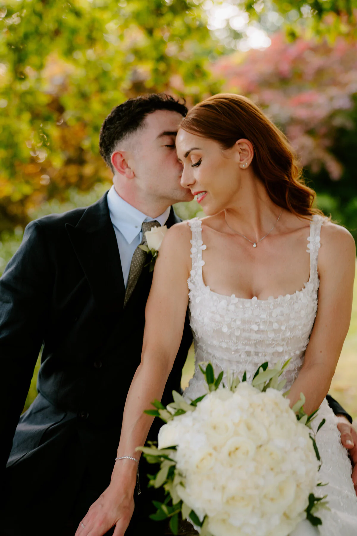 A7r00678 Groom kissing the bride's temple as she holds a white rose bouquet in a floral-applique gown.