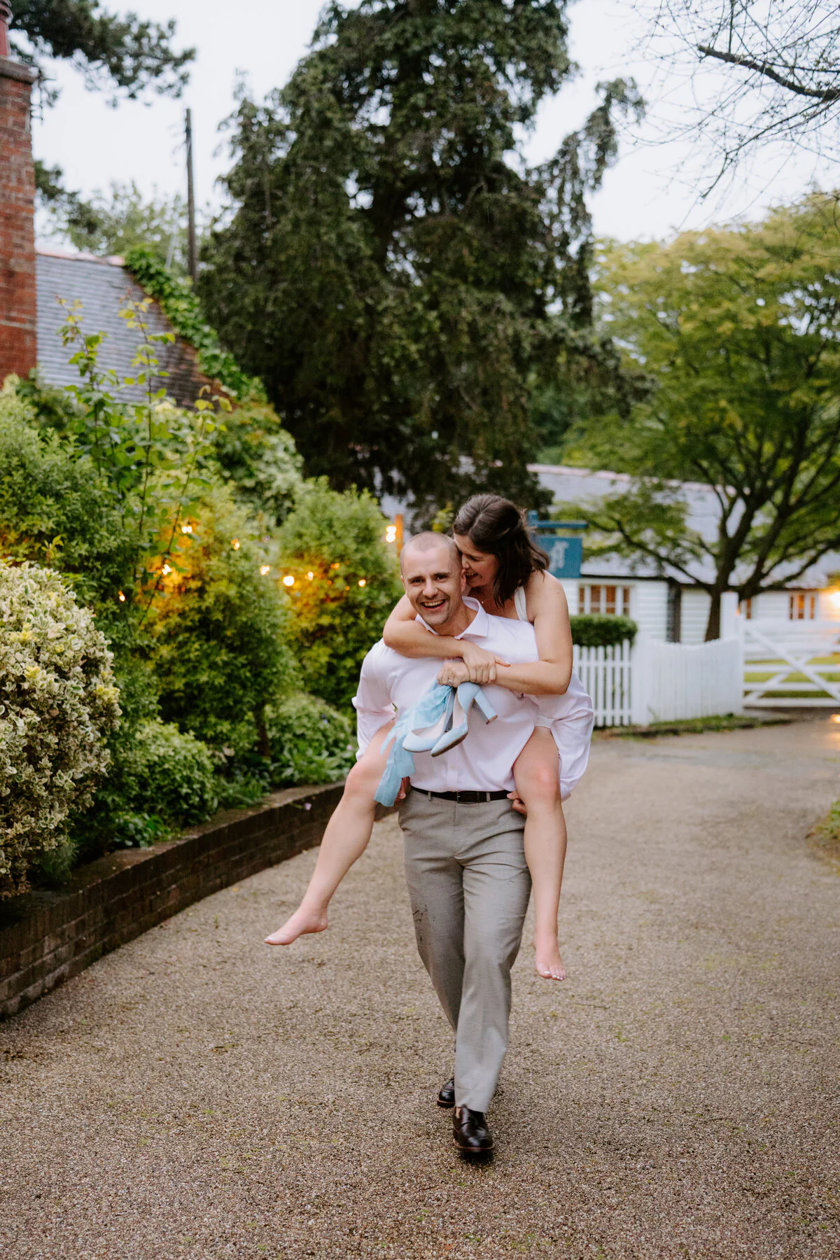Dsc06796 Groom giving the bride a piggyback down a gravel drive past a picket fence.