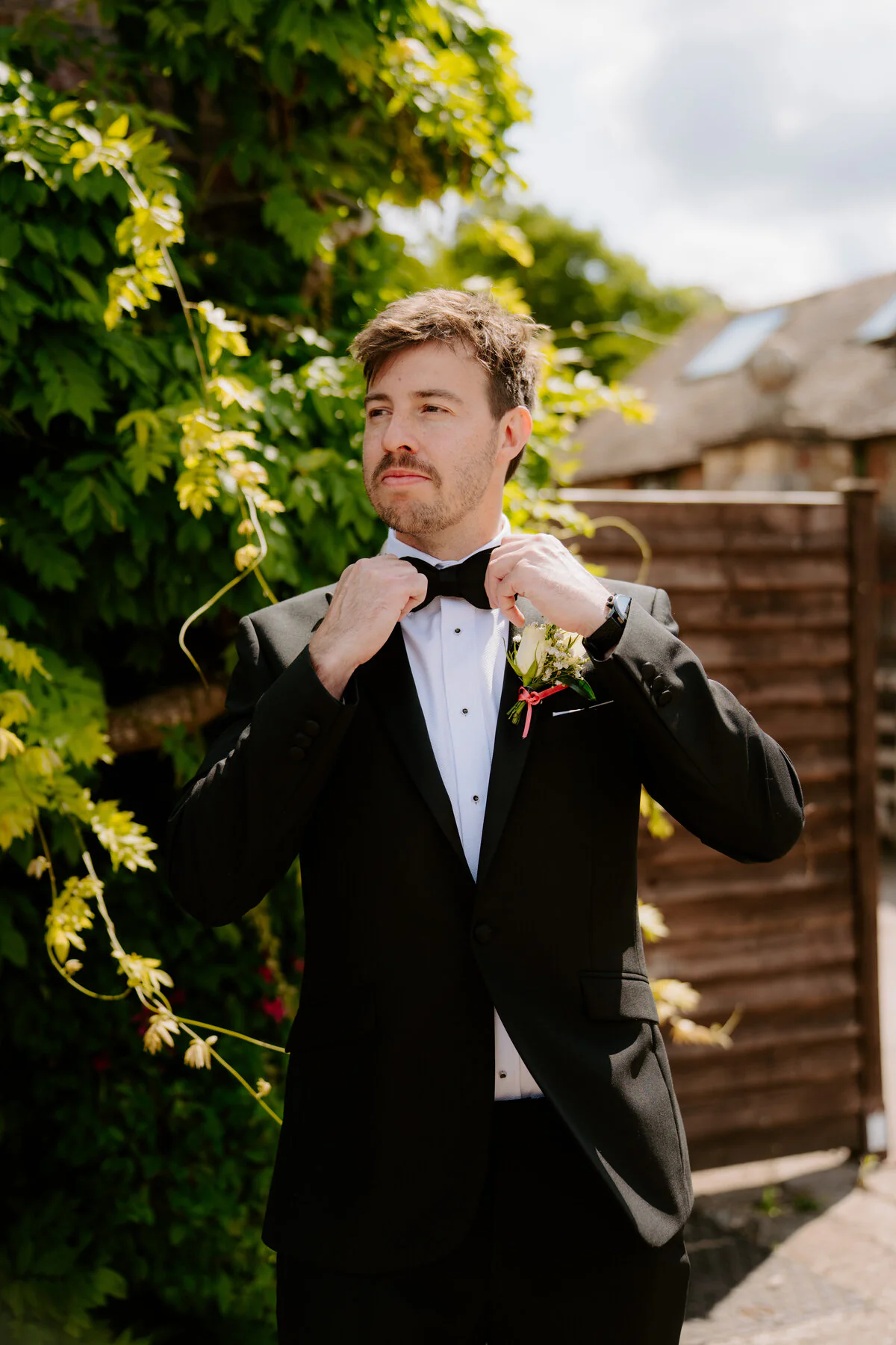 Dsc07392 Groom adjusting his bow tie outside in a black tuxedo with a white rose boutonniere.