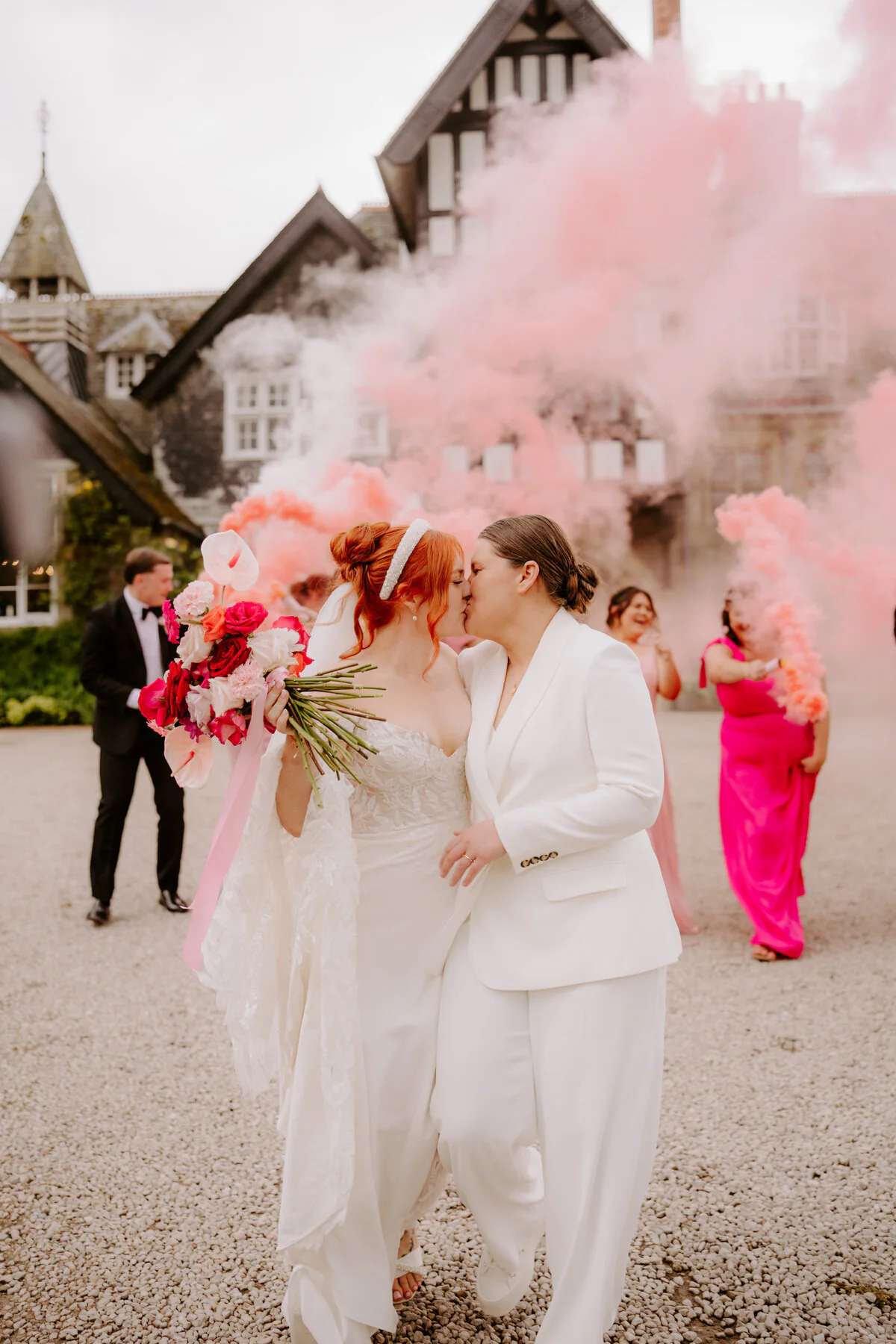 Dsc07011 Two brides kissing behind pink smoke bombs outside a Tudor manor with a red bouquet.