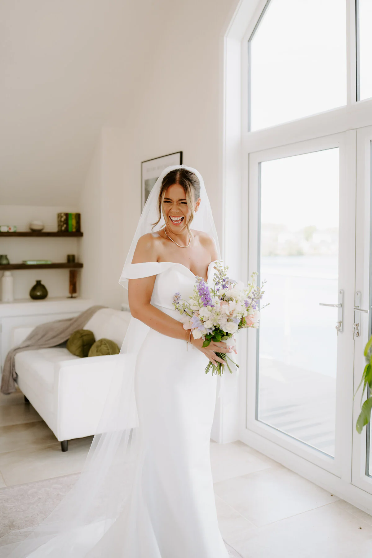 Dsc05794 Laughing bride in an off-shoulder gown with a pastel bouquet in a bright lakeside room.