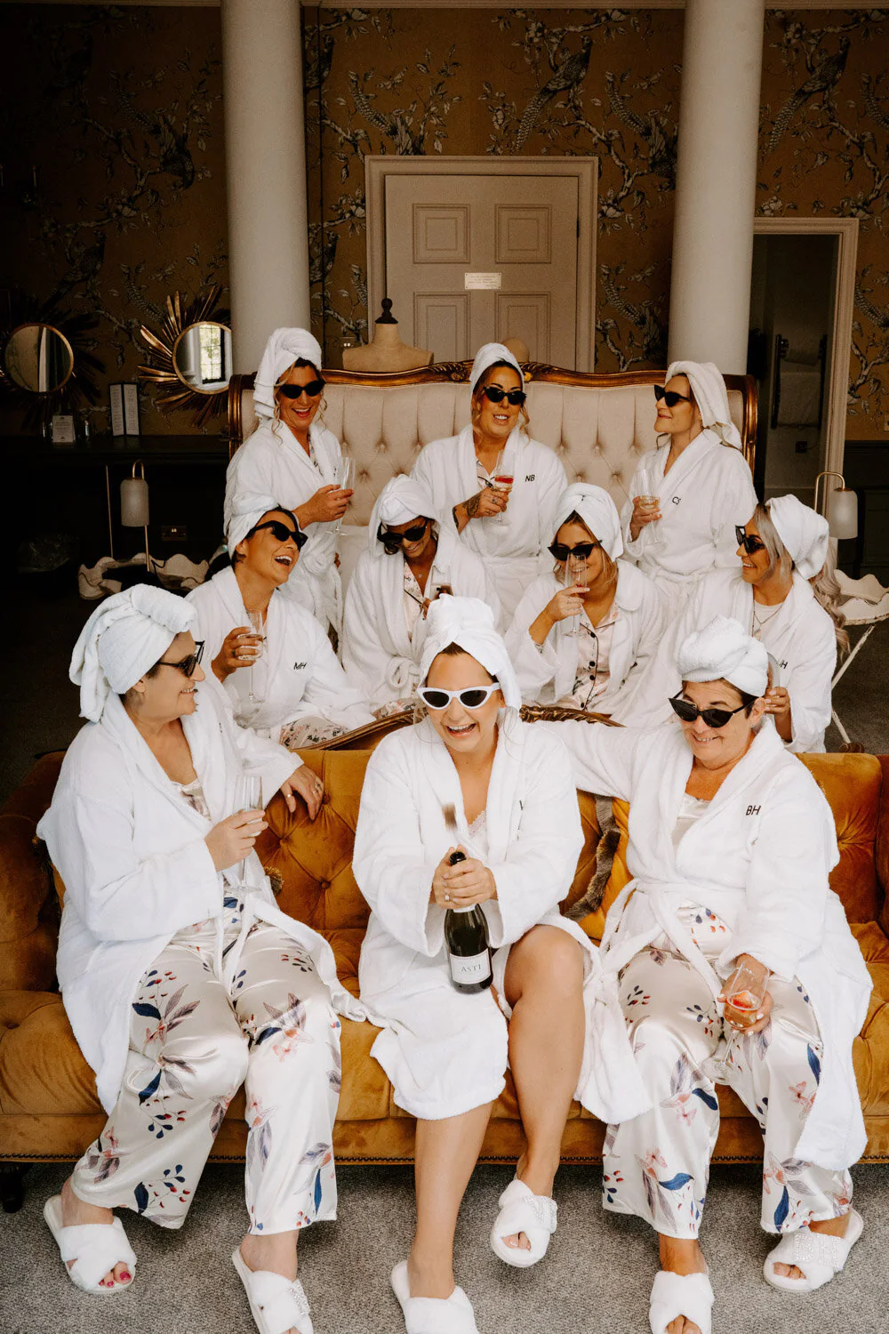 A7308364 6 35 Bridal party in robes with towels on their heads toasting champagne on a velvet sofa.