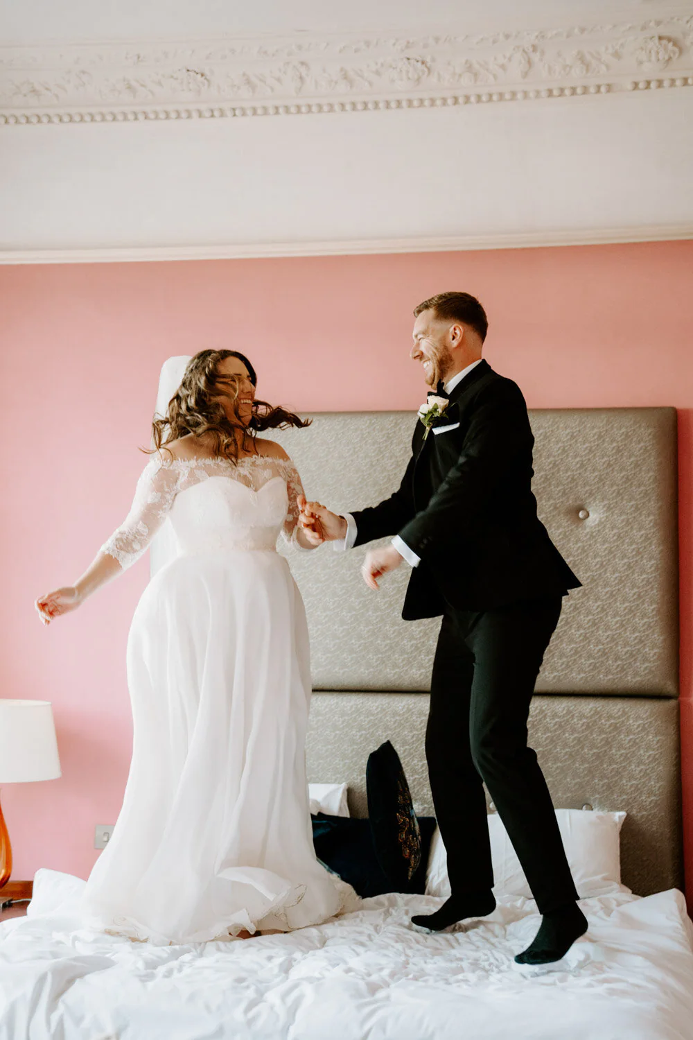 561 Dsc05107 29 Bride and groom jumping on a bed laughing in a pink bedroom.
