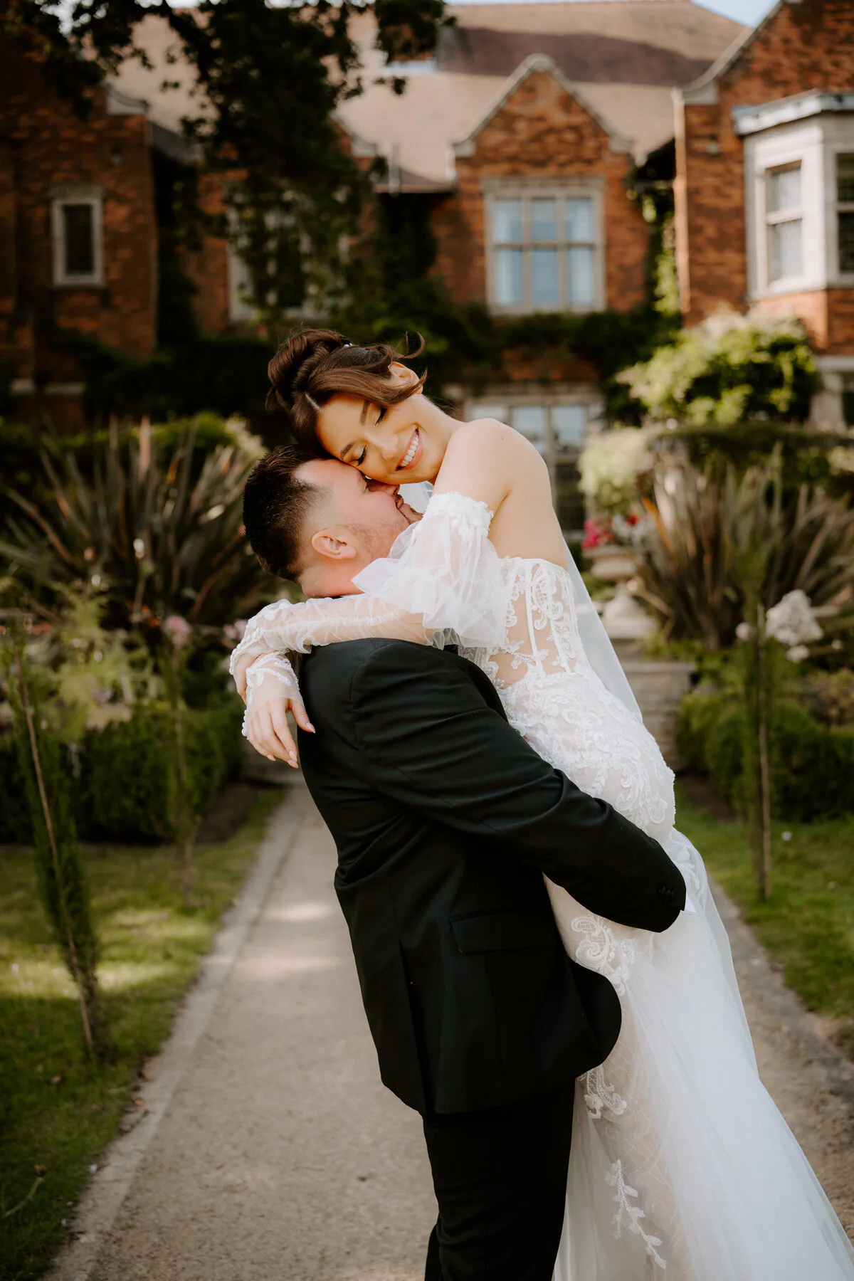 Dsc08823 Groom lifting his laughing bride in an off-shoulder lace gown on a manor garden path.