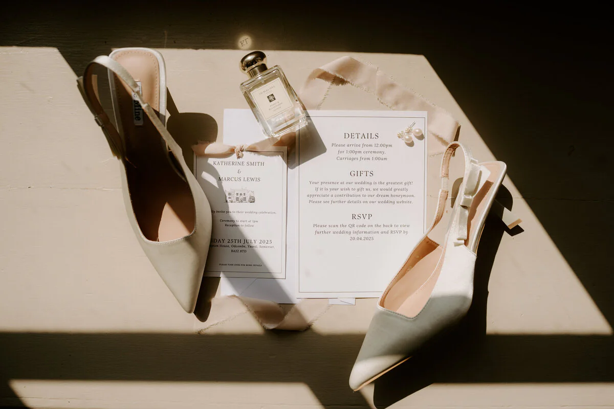 Dsc01241 Flat lay of white satin heels, perfume and Katherine and Marcus's wedding stationery in sunlight.