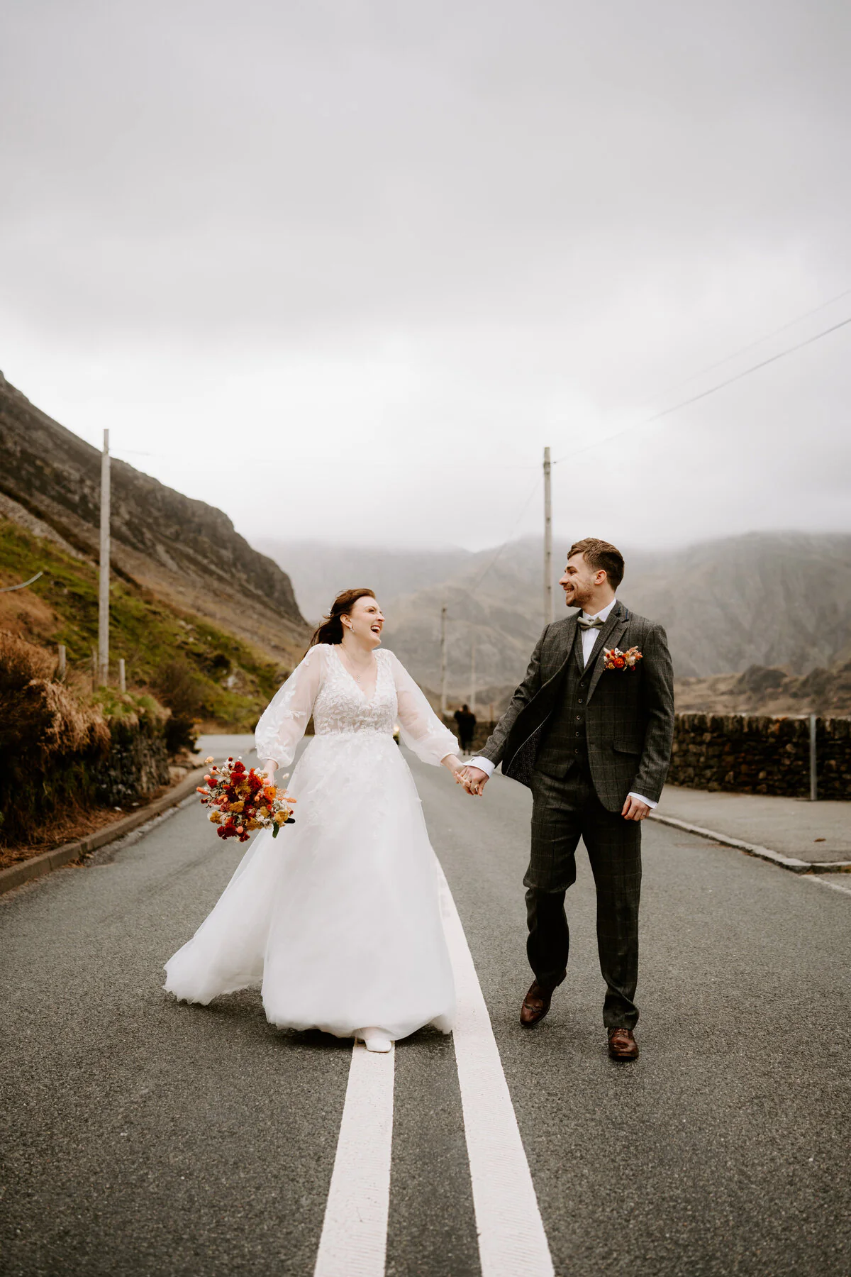Dsc07616 Couple laughing as they walk the centre line of an empty mountain road, with an orange bouquet and tweed three-piece suit.