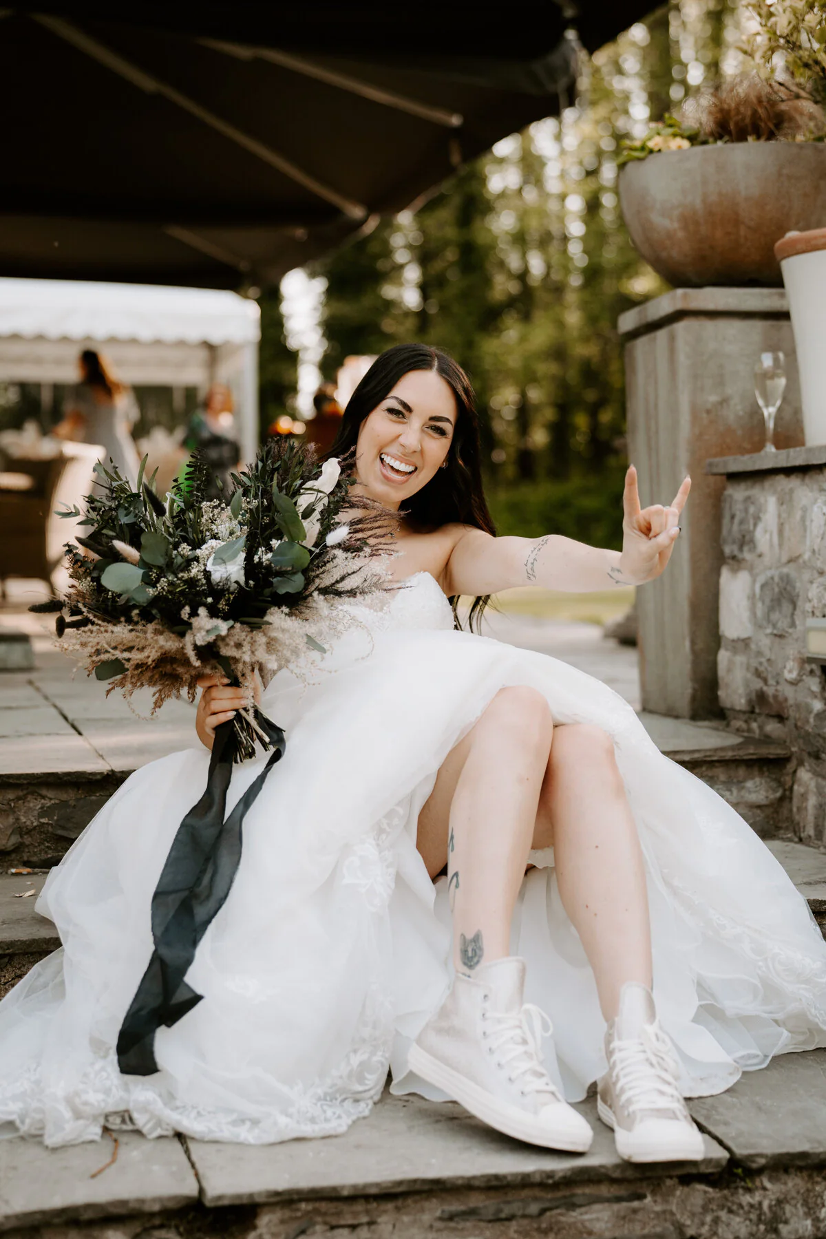 Dsc06600 1 Bride in white Converse trainers flashing a rock-and-roll hand sign on stone steps with a dried bouquet.